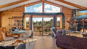 A cozy cabin interior with a pool table, bookshelves, a fireplace, and a large window overlooking a forest.