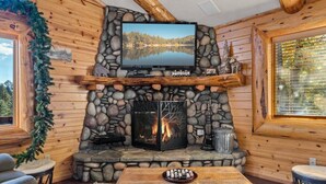 A cozy cabin interior with a stone fireplace, a television mounted above it, and a wooden table with a plate of snacks.