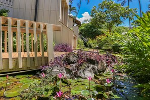 Un jardín tranquilo con una valla de madera, un estanque con flores de loto y abundante vegetación.