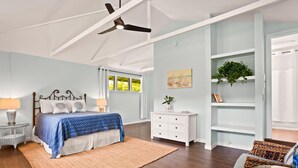 Une chambre à coucher comprenant un lit, des tables de chevet, un ventilateur de plafond, une fenêtre, une plante et une bibliothèque.