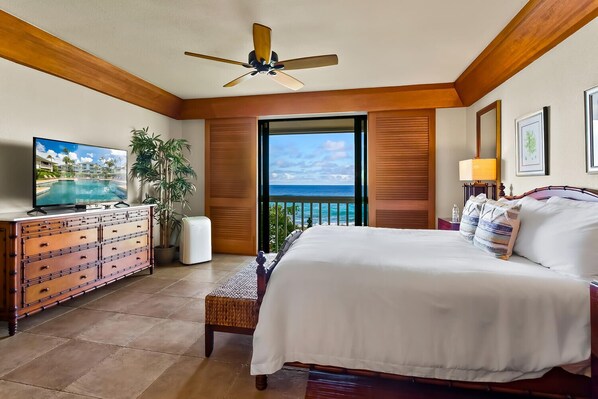 A hotel room with a bed, a TV, a ceiling fan, a view of the ocean, and a wooden floor.