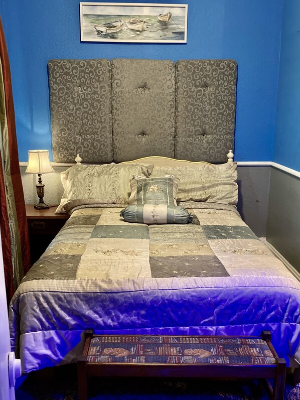 Quarto com parede azul, cabeceira estofada cinza, colcha acolchoada e mesa de cabeceira com abajur.