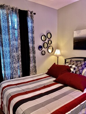 Quarto com cama, cortinas estampadas, espelho fixado na parede, luminária e pintura.