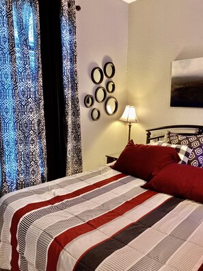 Una cama bien hecha con una colcha a rayas, un espejo en la pared, una lámpara y cortinas estampadas.