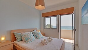 Ein Schlafzimmer mit einem hölzernen Bett, einem Nachttisch, einer Nachttischlampe, einem Fenster mit Meerblick und einem Gemälde an der Wand.