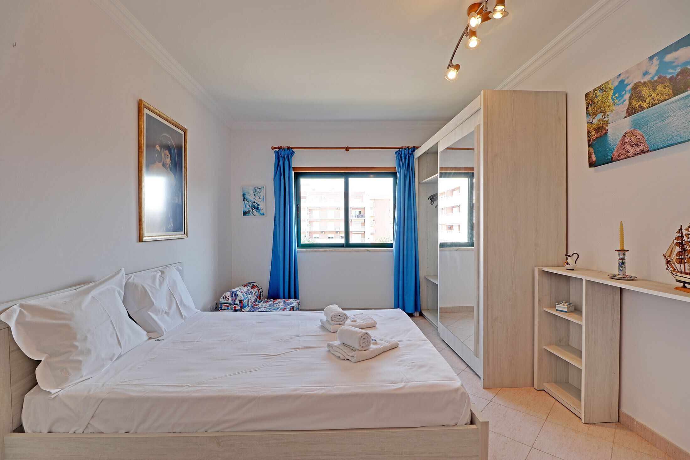 A hotel room with a bed, a window with blue curtains, a painting on the wall, and a small shelf with a candle and a ship model.