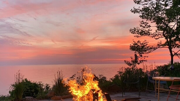 Bocche di fuoco con tronchi ardenti, circondate da un patio lastricato con tavolo e sedie, affacciato su uno specchio d'acqua al tramonto.