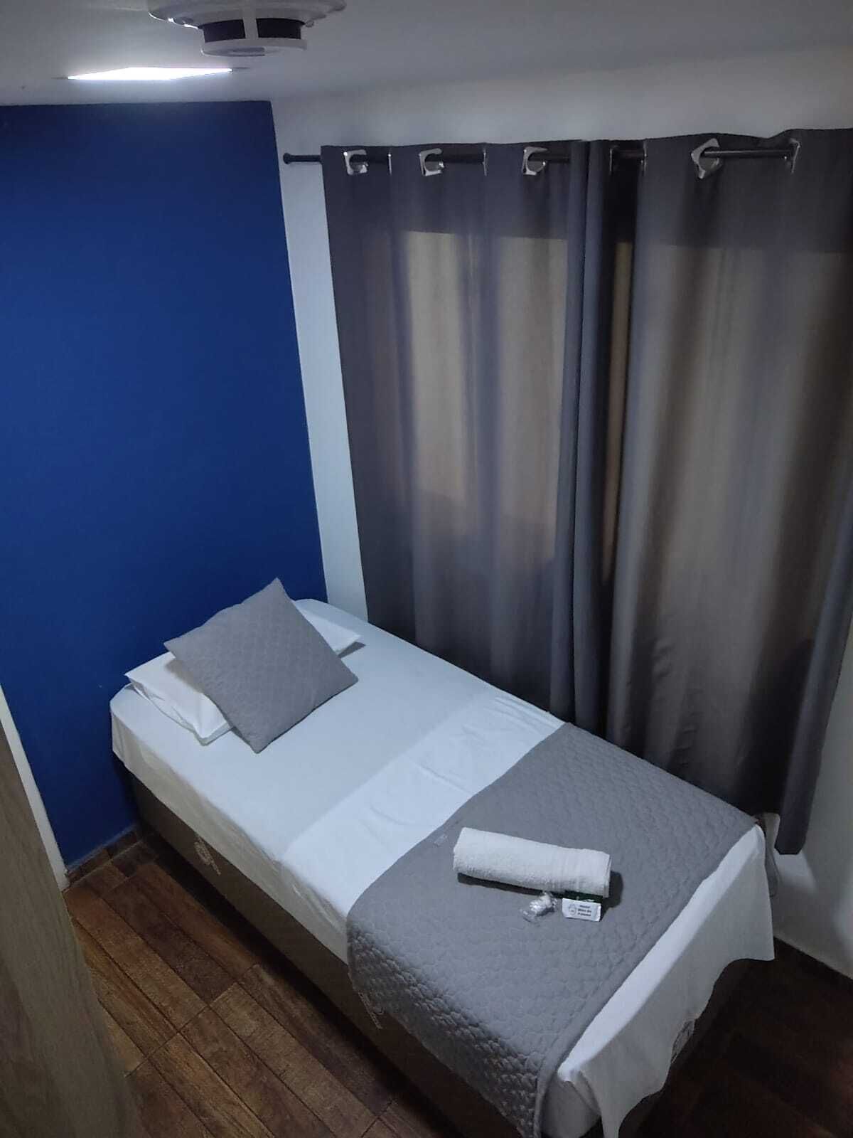 A single bed with a grey blanket and pillow, a white mattress, and a grey quilted cover.