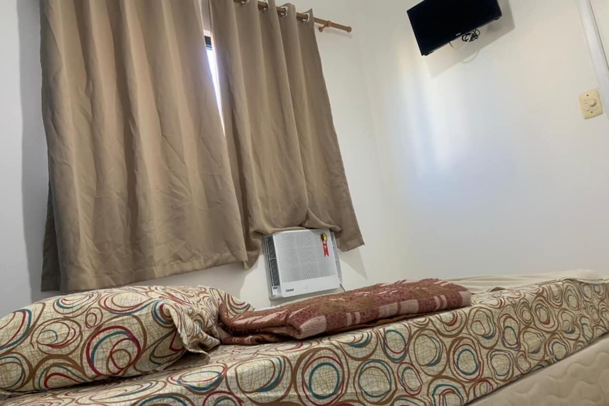 Una habitación con una cama, cortinas color beige, aire acondicionado de pared y un televisor en la pared.