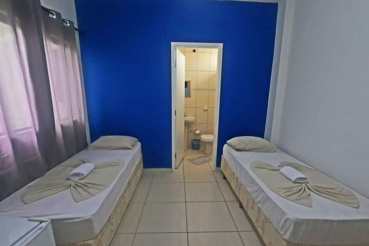 Una habitación con dos camas individuales, un baño con lavamanos e inodoro, y una puerta que da a otra habitación.