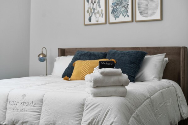 Una cama bien tendida con una colcha blanca, un edredón doblado y una almohada con etiqueta.