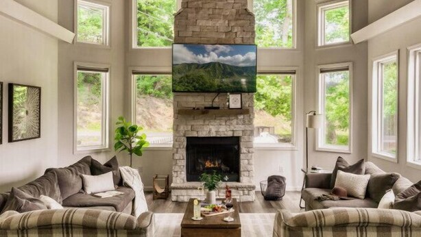 A spacious living room with a stone fireplace, large windows, and a high ceiling with wooden beams. - Hot Tub/Game Room/Theater/Fire Pit/VIEWS/Luxury! - Ski View Lodge (Gatlinburg)