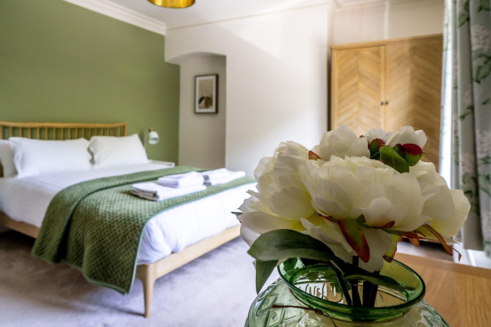 A neatly made bed with a green blanket, a vase with white flowers, and a wooden wardrobe in a hotel room.
