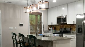 A modern kitchen with white cabinets, a stainless steel refrigerator, a microwave, and a large island with bar stools. - Quiet & Chic 3 BR Cottage w/ Private Garden Near Downtown Asheville (Asheville)