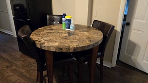 Una mesa de comedor con cubierta de mármol, rodeada por cuatro sillas. Se ve un refrigerador negro al fondo.