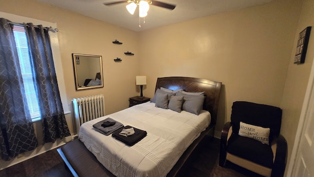 Un dormitorio con cama, ventilador de techo, mesita de noche con lámpara, sillón con cojín y ventana con cortinas.