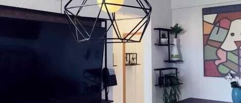 A modern living room with a flat-screen TV, a kitchen area with a dining table, and a geometric pendant light fixture.
