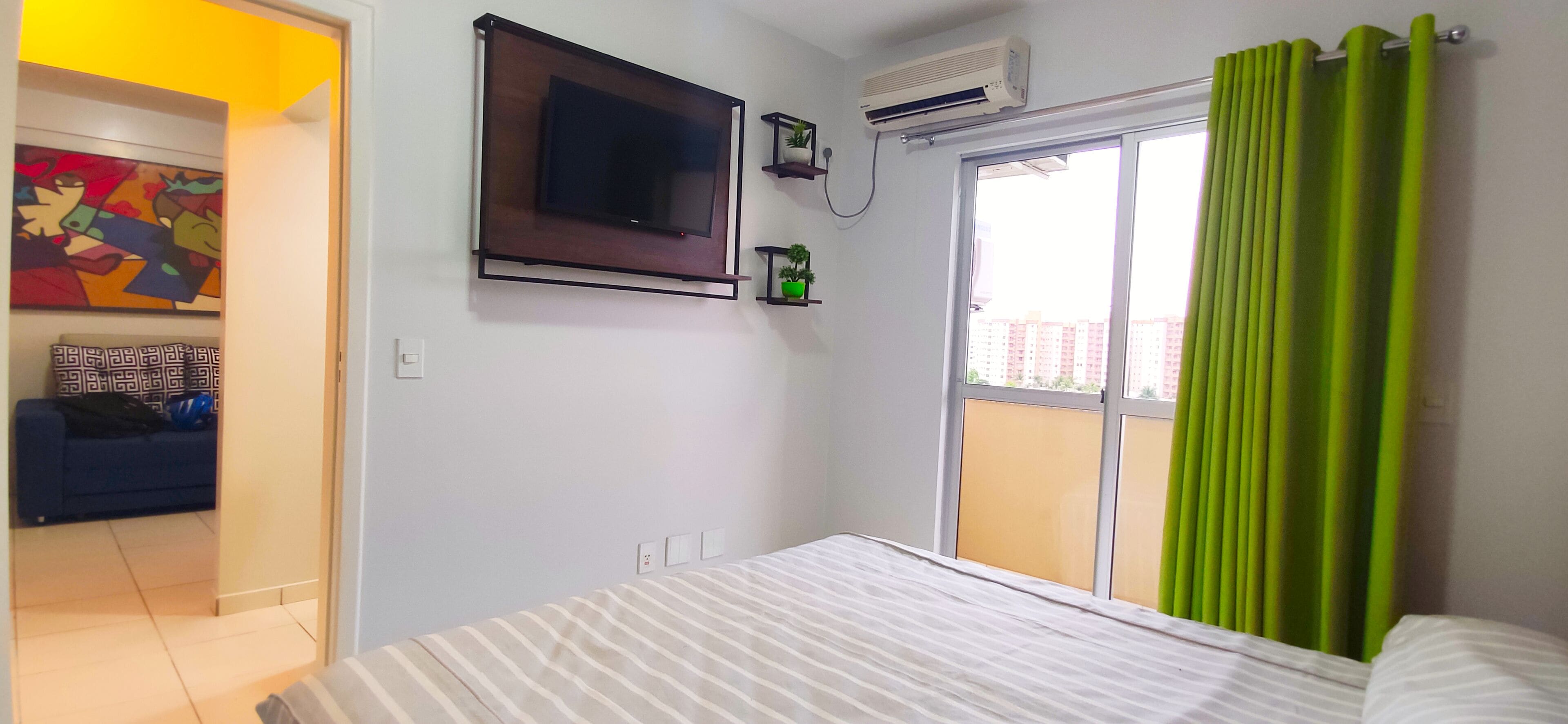 Quarto com cama, TV na parede, ar condicionado, janela com cortinas verdes e um quadro colorido na parede.
