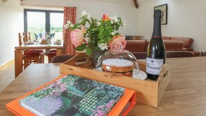 A wooden table with a bottle of Prosecco, a vase with flowers, a box with a book, and a view of a dining area with a window. - Swallows Nest (Sturminster Newton)