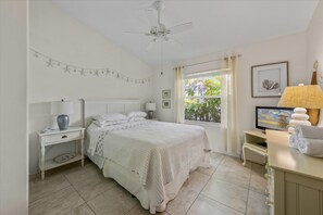 Un dormitorio con cama, mesitas de noche, ventilador de techo, ventana con cortinas y televisor.