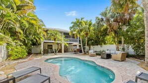 A pool area with a curved edge, surrounded by patio furniture and palm trees. - Isle Relax: Close to Gulf of Mexico and Pine Ave. (Anna Maria)