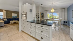 A modern kitchen with white cabinetry, a central island, and a dining area with a table and chairs. - Isle Relax: Close to Gulf of Mexico and Pine Ave. (Anna Maria)