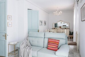 Un canapé bleu clair agrémenté d’un coussin coloré, une porte blanche et un lustre dans une pièce aux murs blancs.