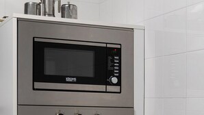 Une cuisine compacte dotée d’un four à micro-ondes et d’un four encastrés, d’une hotte de cuisine en acier inoxydable et d’un plan de travail blanc.