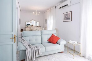 Une pièce avec un canapé bleu clair, un coussin rouge, une table d’appoint à cadre doré et un appareil de climatisation blanc fixé au mur.