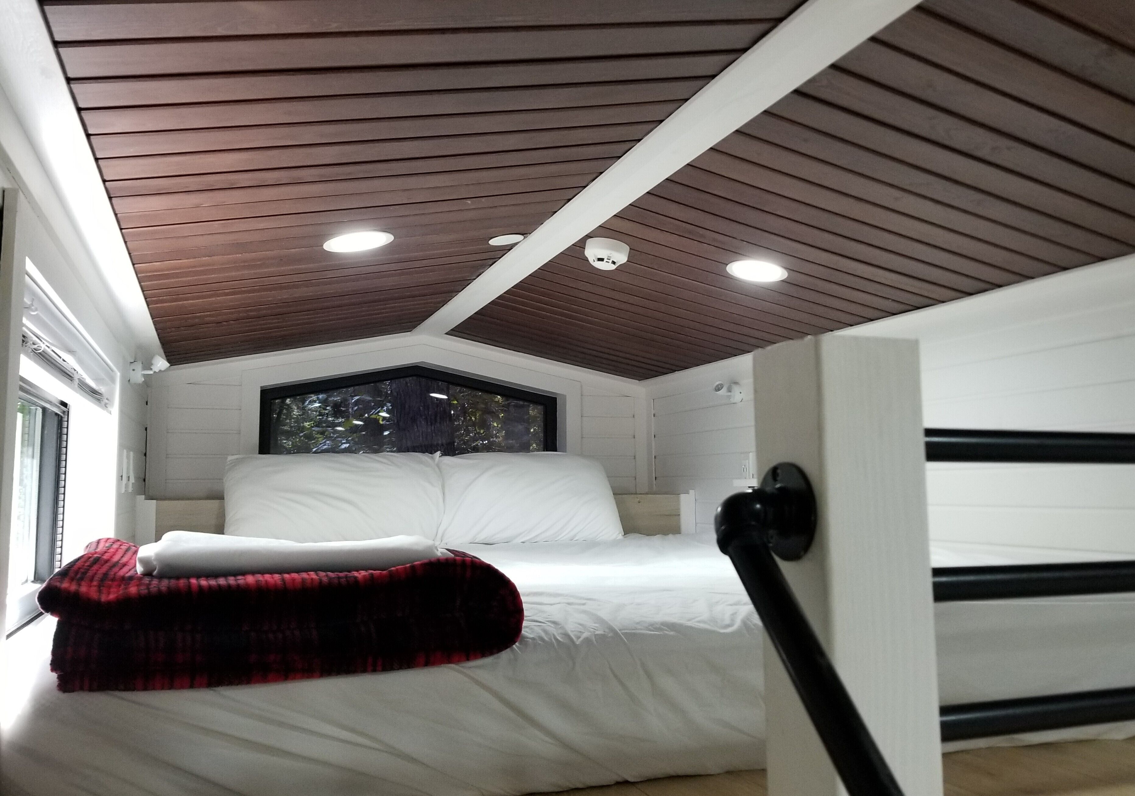 A bedroom with a wooden ceiling, white walls, a bed with white bedding, a red and black plaid blanket, and a window with a view of trees.
