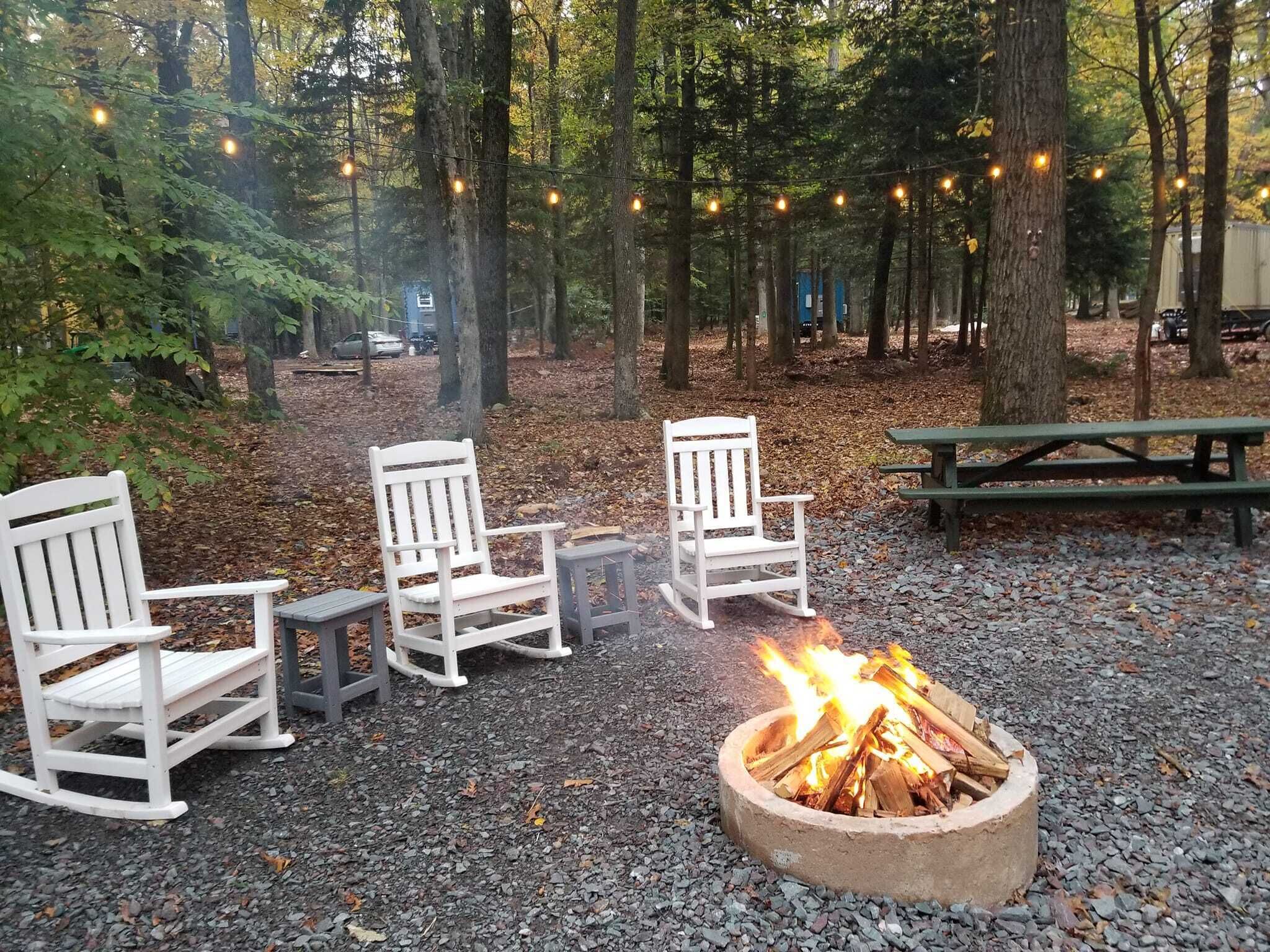 A campsite with three white Adirondack chairs, a small table, and a fire pit with logs.