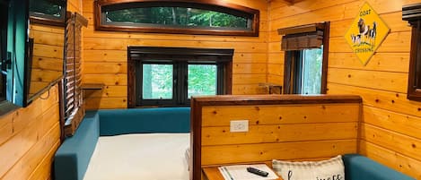 A cabin with wooden walls, a bunk bed, a desk, and a window with a view of trees.
