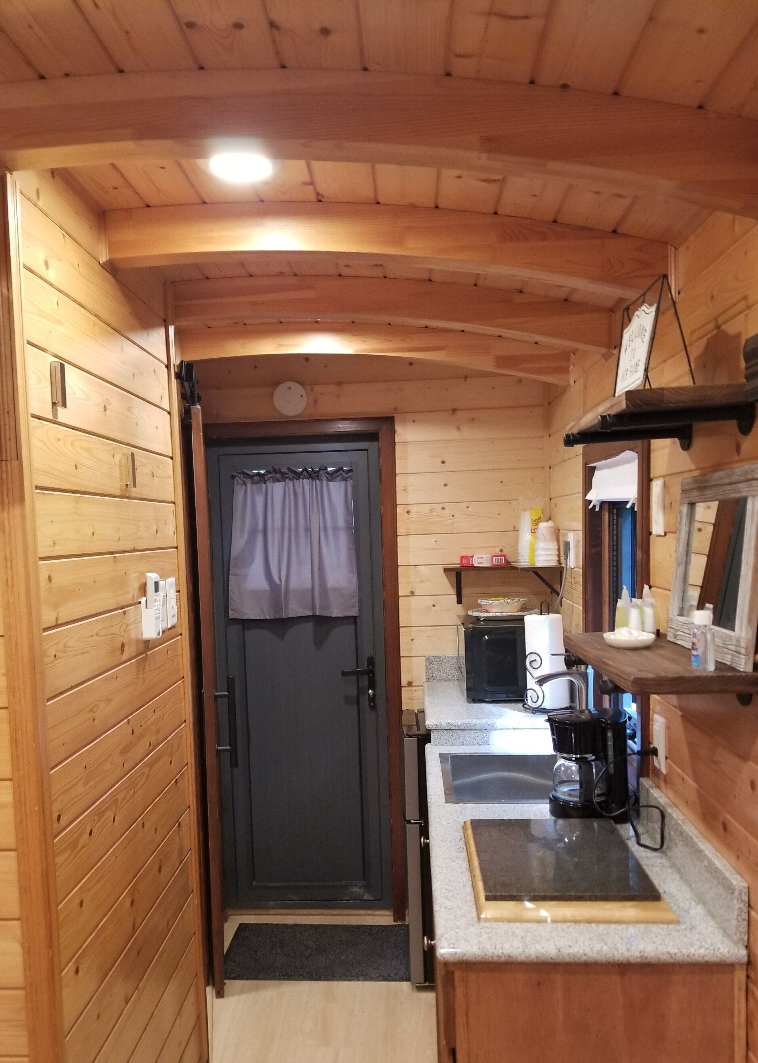A compact kitchen with wooden walls, a door with a curtain, a microwave, a coffee maker, and a small shelf with items.