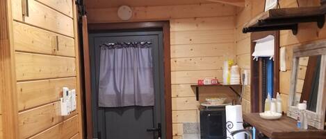 A compact kitchen with wooden walls, a door with a curtain, a microwave, a coffee maker, and a small shelf with items.