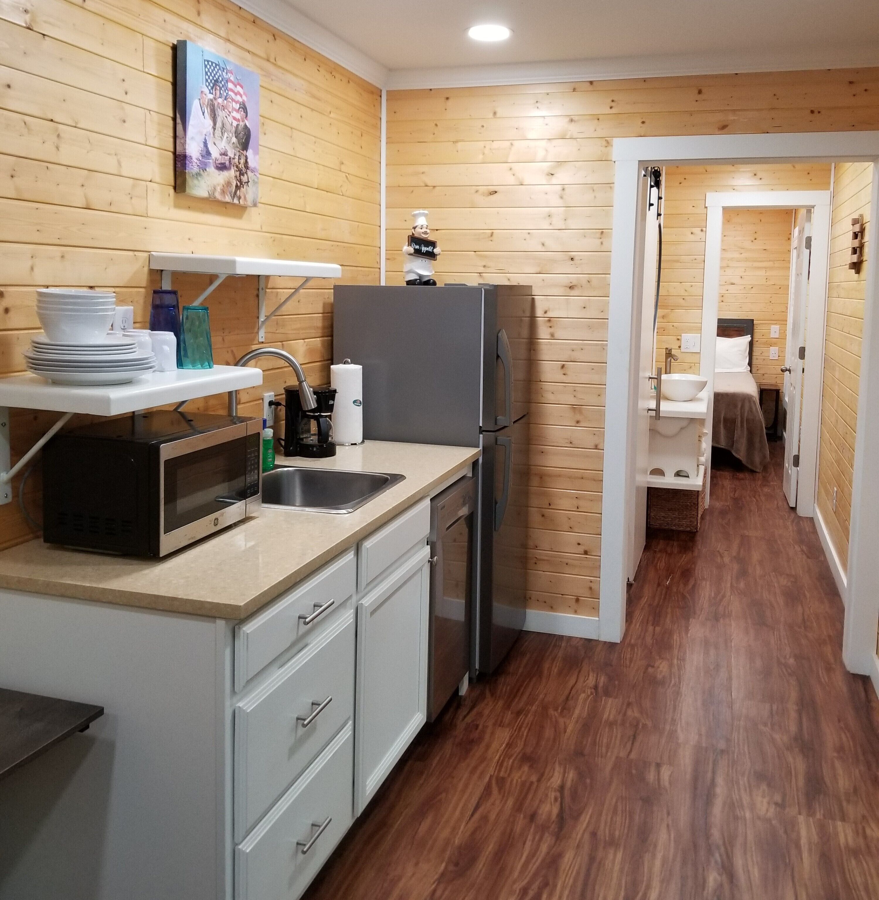 A compact kitchen with a microwave, sink, and refrigerator. There is a small dining area with a table and chairs, and a hallway leading to a bedroom.