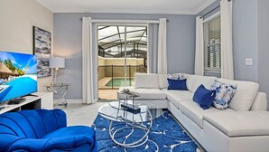 A modern living room with a white sofa, blue armchair, and a glass coffee table. A flat-screen TV is on a stand, and there is a glass door leading to a balcony with a pool. - EV_2861 - 4BR / 3BA Storey Lake Townhome near to Disney, Private Pool & Resort (Kissimmee)