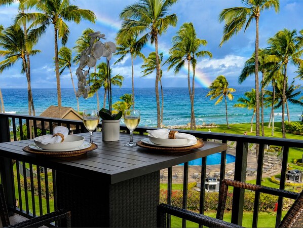 A table set with white plates, bowls, and wine glasses, overlooking a beach with palm trees and a view of the ocean.