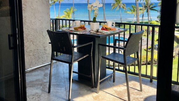 A balcony with a table, chairs, and a view of the ocean and palm trees.