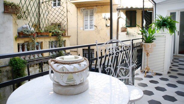 A balcony with a white table and chairs, potted plants, and a metal railing. - Luxury apartment residence "La Fontana" (Lucca)