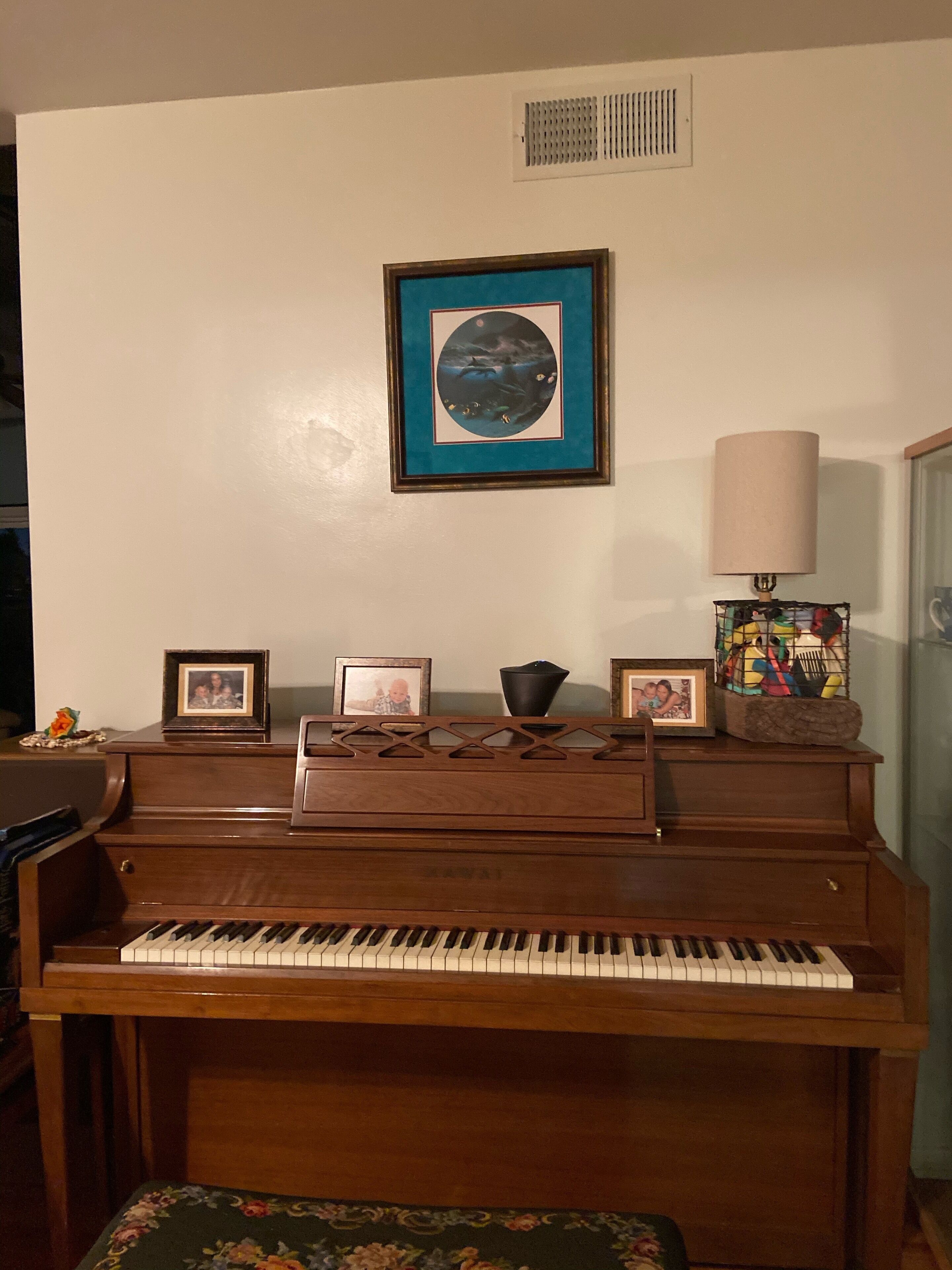 Un piano sur lequel sont posées trois photos encadrées, une lampe et une bouche d’aération au mur.