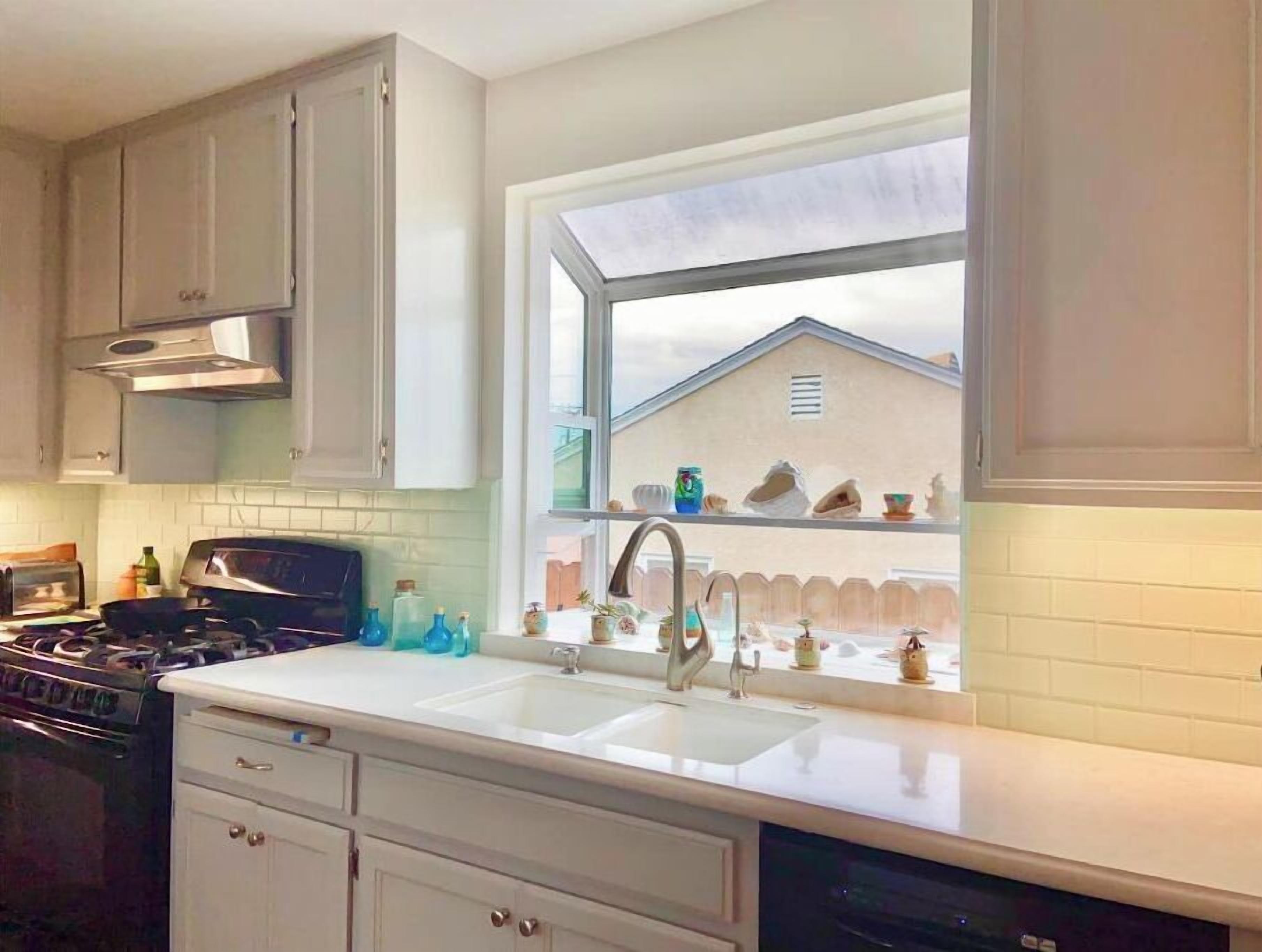 A kitchen with white cabinets, a stove, a sink, and a window overlooking a house.