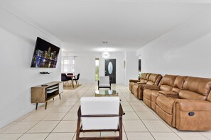 A spacious living room with a large leather sofa, a white armchair, a glass-top coffee table, and a flat-screen TV mounted on the wall.