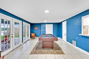 Uma mesa de bilhar numa sala com paredes azuis e detalhes brancos, um tapete de padrão de leopardo e uma porta que dá para o exterior.