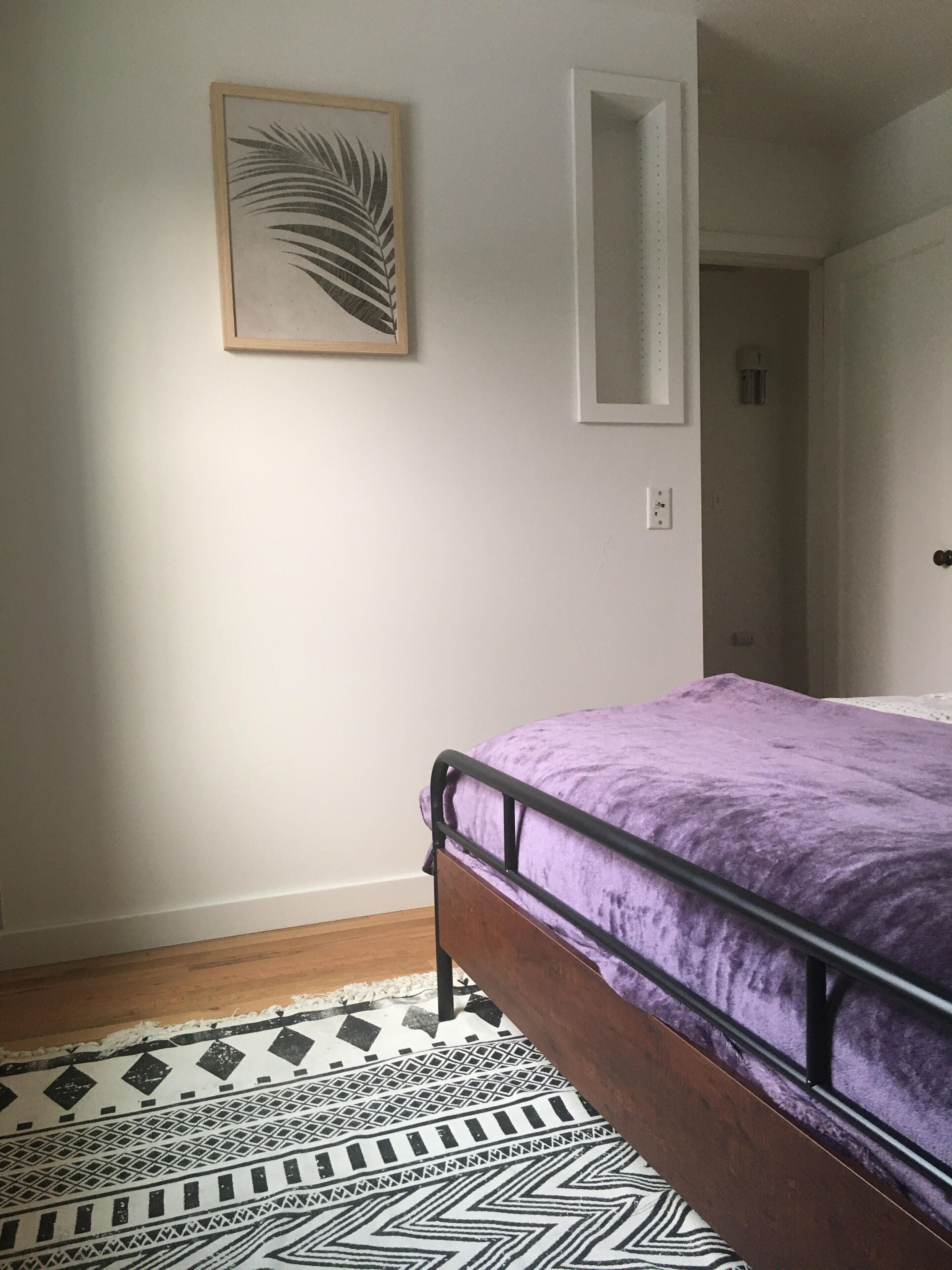 A bedroom with a bed, a patterned rug, a framed artwork, and a door leading to another room.