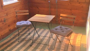 A small, cozy room with wooden walls, a striped rug, a table, two chairs, and two windows. - Oak Street Farm House (Omak)
