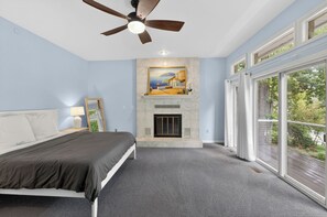 A bedroom with a bed, a ceiling fan, a fireplace, and large windows.
