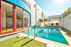 Une maison moderne dotée d’une piscine, de grandes fenêtres et d’un espace extérieur aménagé avec un coin salon couvert.