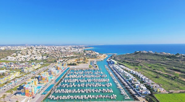 Un port de plaisance situé sur la côte, avec de nombreux bateaux amarrés, un bâtiment abritant les services du port et un quartier résidentiel aux bâtiments aux couleurs vives.
