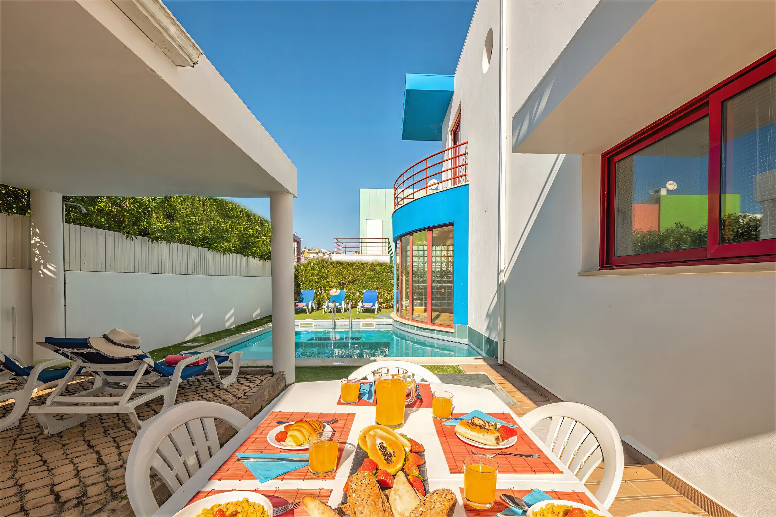 Ein Essbereich am Pool mit Tisch, gedecktem Frühstück, Liegestühlen und Blick auf Pool und Gebäude.