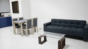A modern living room with a glass coffee table, dark blue sofa, and a dining area with a blue table and chairs.